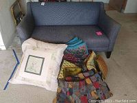 Blue denim fabric love seat with decorative and floor pillows stacked in front of it on carpeted floor.