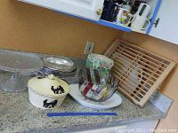 Photo of kitchen counter showing glass cake stands, ceramic cookie jar with lid and animal silhouettes, metal striped mixing bowl, clear glass mixing bowl with a fabric cover, wooden drying rack, and wooden cutting boards