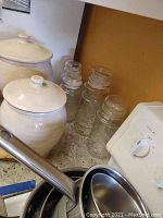 Two large white ceramic canisters with lids placed on a kitchen countertop next to four clear glass storage jars with lids. Stainless steel ladle handle partially visible in foreground.