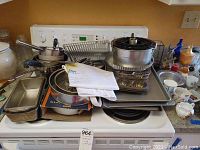 Photo showing assorted metal baking pans, muffin trays, cookie sheets, cooling rack; several pots and frying pans stacked; white papers possibly packing instructions or recipe sheets