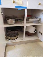 Upper shelf with stacked metal mixing bowls, rectangular baking pans, and other kitchen items inside a white cabinet