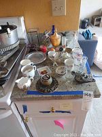 Kitchen countertop filled with various home decor items including figurines, tea cups, metal dishes, stone carvings, and bottles with cork tops.
