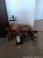 Full view of all copper and metal items including fondue pot, kettle, pitcher, and bookends arranged on carpet.