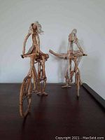 Two handcrafted wooden bicycle rider figures wrapped in rope, showing detailed bicycles and figures.