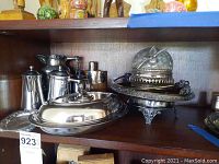 Photo showing assortment of silver plated tea set and serving dishware on a shelf including teapot, flask, covered dish, and lidded bowl.