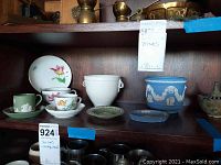 Shelf view showing multiple teacups and saucers with floral and classical Wedgwood style, white handled urn-shaped cup, and blue jasperware pottery pieces
