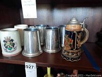Shelf showing 4 metal beer steins with clear bottoms, 1 pewter beer stein, and 2 ceramic beer steins including one with colorful scenes and floral borders.