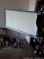 Projector screen mounted on a tripod with visible missing portion on one side, overall white with blue edging.