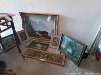 Six framed art pieces grouped on the floor against a wall showing variety in size, frame style, and condition. One large painting has a worn, damaged frame.