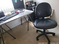 View of the office desk with computer monitor, keyboard, and mouse on top; black office chair positioned next to it.