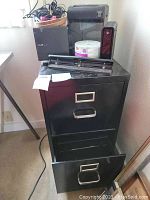 Photo showing top and front of black two-drawer metal file cabinet with one drawer open, black 3 hole punch on top