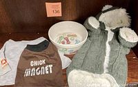 Lot of dog clothing and ceramic dog bowl shown on wooden background including army green sweater, brown T-shirt, gray sweatshirt, and decorative dog bowl.