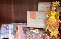Photo of the doll, original box, certificate, and various magazines arranged on a surface