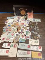 Photo of the contents of a Punch cigar box showing an assortment of vintage stamps on paper, various old coins, vintage envelopes with stamps, a currency note, and a pink tag numbered 23.