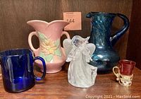 Collection of vintage blue glass and other collectibles including pink floral vase, blue pitcher, cobalt mug, clear glass angel figurine, and small shot glasses.