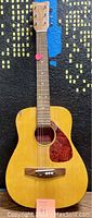 Front view of Yamaha FG-Junior acoustic guitar on black and blue carpet background, guitar pick visible on strings near headstock.