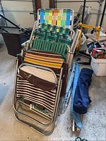 Three stacked aluminum folding beach chairs with woven seats in green and yellow, with a vinyl striped lounge chair on top, all leaning against the garage wall with other items around them.