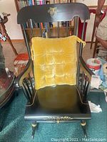 Full view of the black wooden rocking chair with yellow cushion, set on carpeted floor near bookshelves and other furniture.
