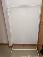 Full view of a white metal clothes rack with rectangular base and curved feet against a wall