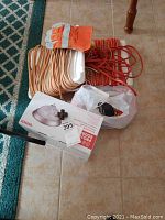 Photo showing coiled beige and orange outdoor extension cords, orange and white work gloves, and light fixture box, on tiled floor near turquoise rug.