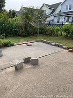 Wide view showing grey concrete border blocks arranged on a concrete patio with red round and square stepping stones in the background along garden edge, including lawn and surrounding houses.