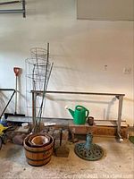 Front view of garden supplies this includes metal tomato stakes and cages, green watering can, wooden barrel planters, garden bricks, metal umbrella stand, shovel, and more items arranged against a garage wall