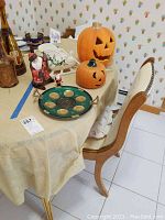 Photo showing a beige table with a large carved ceramic Halloween pumpkin, a smaller pumpkin, two Santa Claus figurines (one sitting, one standing), and a green metal round Seder plate with gold accents and inscriptions.