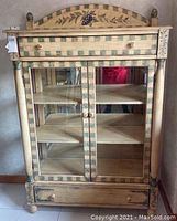Front view of hand painted wooden kitchen cabinet showing double glass doors, drawer above and below, grapevine motif on arched top, and checkered border design.