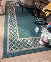 Full view of large green area rug with beige lattice-patterned border; visible wear and creases.