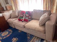Beige upholstered three-seat sofa with soft cushions and two matching pillows, placed on a carpet with dolls on seat.