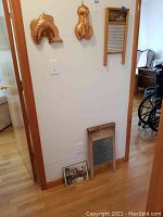 Photo showing two copper molds hanging on the wall, a wooden-framed washboard with ceramic trough and ridges, a wooden-framed washboard with metal scrubbing surface leaning against the wall and a rustic framed painting on the floor.