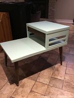 Side angled view showing two-level off-white painted end table with slotted side panel and tapered legs.