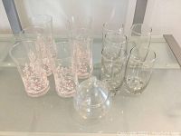 Photo showing six tall drinking glasses with pink floral design and five plain drinking glasses plus one clear glass dish with lid all displayed on clear shelving.