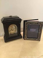 Photo showing the dark wood mantle clock with glass front and brass mechanism beside the silver-plated photo album with detailed border frame, both standing upright against a plain background.