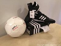 Photo showing a pair of new black Adidas men's soccer shoes size 6.5 with white stripes displayed on top of the original Adidas box alongside a white promotional soccer ball with 'BEND IT like BECKHAM' text.