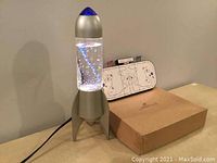 Photo showing a silver rocket-shaped water tornado lamp with swirling illuminated water, and next to it a wooden cribbage board with a hockey rink printed design.