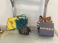 Photo of five different purses including a yellow rectangular purse, turquoise floral purse, black spiral floral purse, clear handbag, and multi-color floral fabric purse with an Eaton's box.