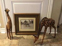 Photo showing two wooden giraffe sculptures and a framed elephant print on a tiled floor in front of a white door.