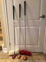 Full view of three golf clubs with persimmon wood heads and two head covers on floor against door.