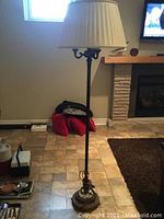 Photo showing the full 3-light floor lamp with pleated white shade, black metal stand and base, functioning in a home setting near a fireplace.