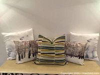 Photo showing five decorative pillows arranged in two rows: two white pillows with polar bear images at each end, two black and white scenic tree design pillows next to them, and a multicolored striped pillow centered in front.