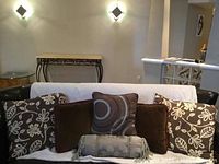 Six brown decorative pillows arranged on a black couch with white towel or throw behind. Two floral pattern pillows on sides, two solid brown pillows, one circle pattern pillow, and one neck roll pillow in front.