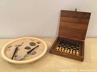 Photo of a round beige dish filled with sand, some smooth stones, and two small wooden tools for a Zen garden next to a square walnut wood case with a magnetic chess set inside.