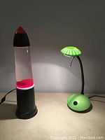 Red lava lamp and green desk lamp side by side, both plugged in and lit up with the lava lamp showing red wax blobs moving inside the clear liquid.