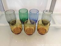 Photo of seven vintage drinking glasses including four tall glasses in grey, green, blue, and light grey, and three shorter amber/gold glasses.