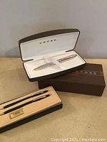 Two separate Cross pen and pencil sets with original boxes displayed on a surface. One set in silver and gold-tone, the other in black and silver-tone.