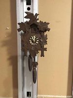 Full view of the wooden cuckoo clock hanging, showing carved leaf and bird details, clock face, pendulum, and pinecone weights.
