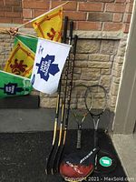 Photo showing three hockey sticks with wooden blades, two Dunlop tennis racquets, two flags (yellow with red lion and white with Toronto Maple Leafs logo), and two Dunlop tennis ball containers on asphalt beside a brick and stone wall.