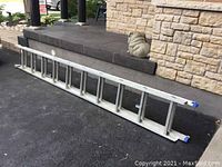 Aluminum expandable ladder laid flat on a blacktop surface near a stone wall and steps outside a building.