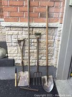 Photo of five used garden tools against brick and stone wall including two shovels, garden fork, garden hoe, and hand fork.
